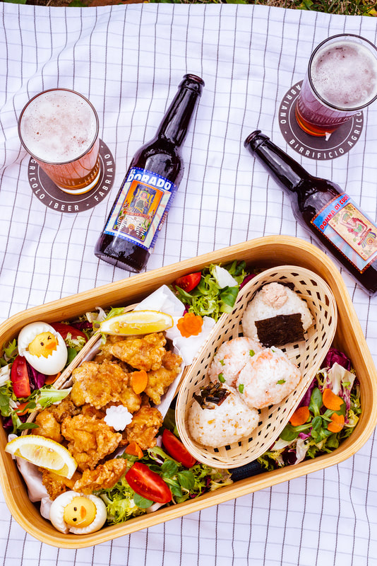 Large bento box lain on a picnic mat accompanied by two bottles of Italian craft beer.