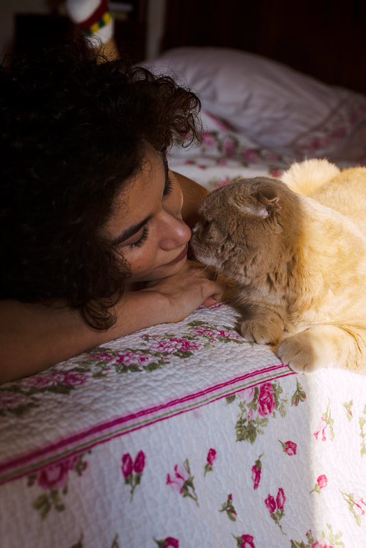 Scottish fold cat kissing a curly haired woman on the bed with floral covers.